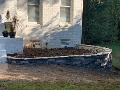Curved stone garden bed beside a white brick house with potted plants and mulch around a window.