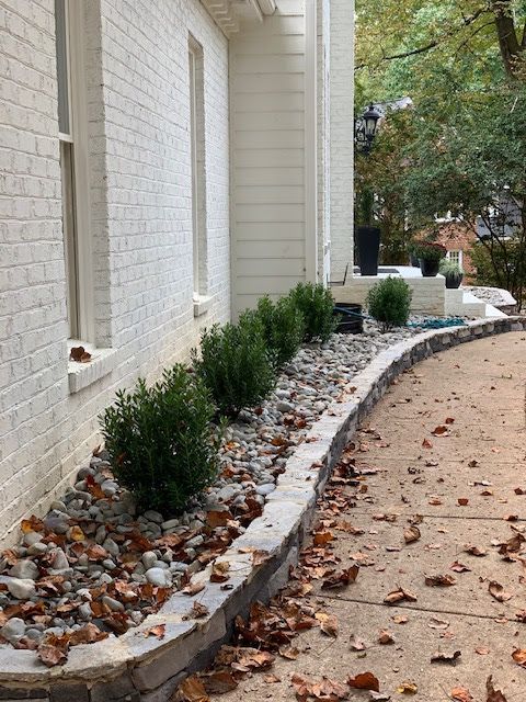 White brick house exterior with a curved stone planter bed and small shrubs along a sandy walkway