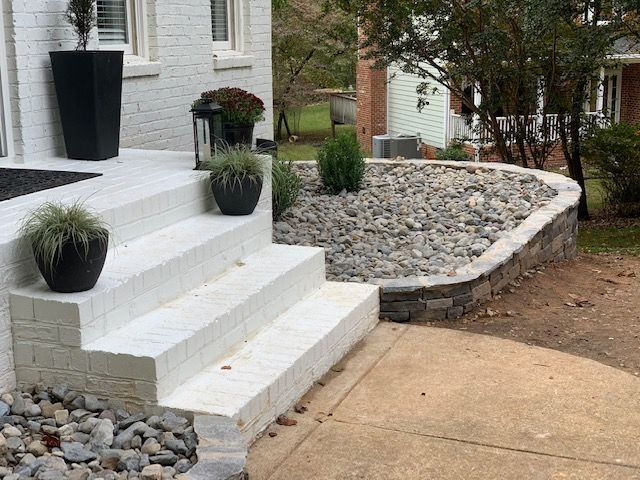 White front steps with potted plants beside a rock-covered garden bed and sidewalk.