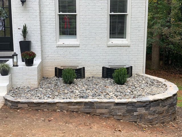 Front of white brick house with curved stone planter bed and potted shrubs inside.