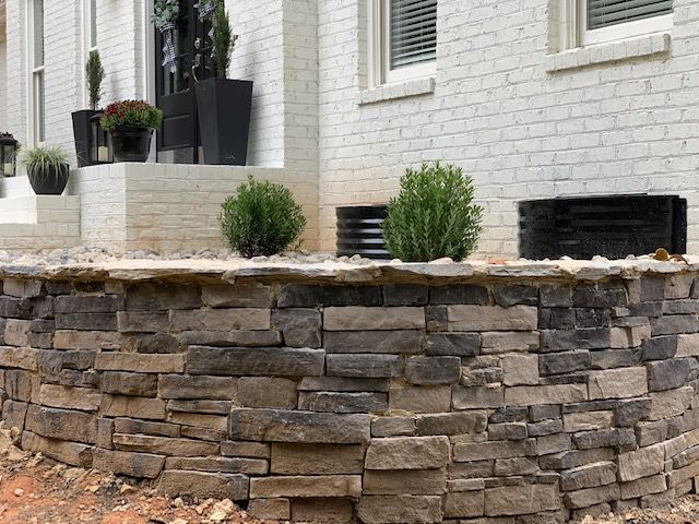 Stone retaining wall with small shrubs in front of a white brick house