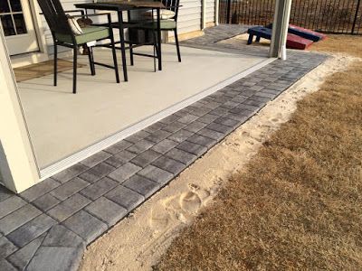 Patio with black table and chairs beside a gray paver path and brown grass, viewed from a porch.
