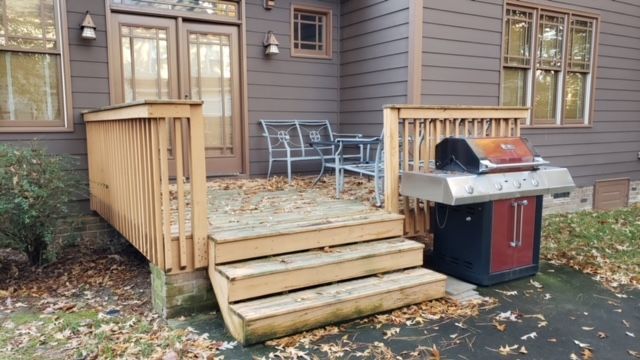 Wooden deck steps leading to a patio with table and chairs beside a red grill by a house