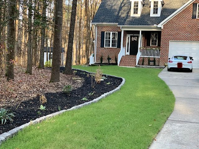 Front yard with a curved stone edge, green lawn, driveway, and a brick house with a white car in front.