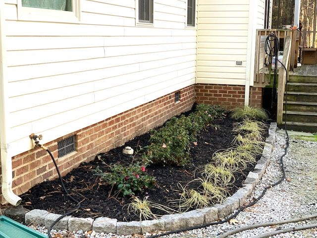 Mulched garden bed along house foundation with brick border, hose faucet, and stairs on the right