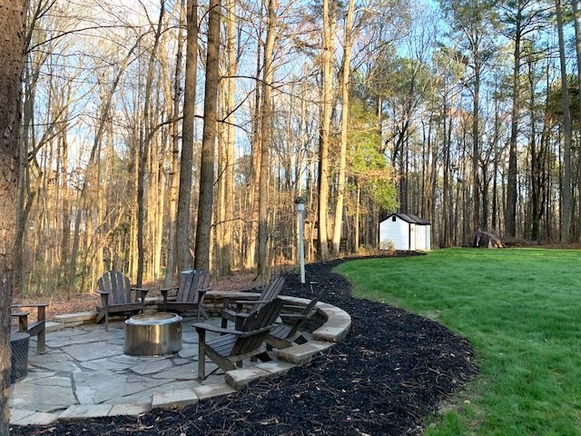 Stone fire pit and curved wooden benches in a wooded backyard, with a small white shed in the background.