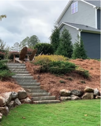 Stone steps lead up a landscaped hillside beside a gray house with outdoor chairs on the landing.