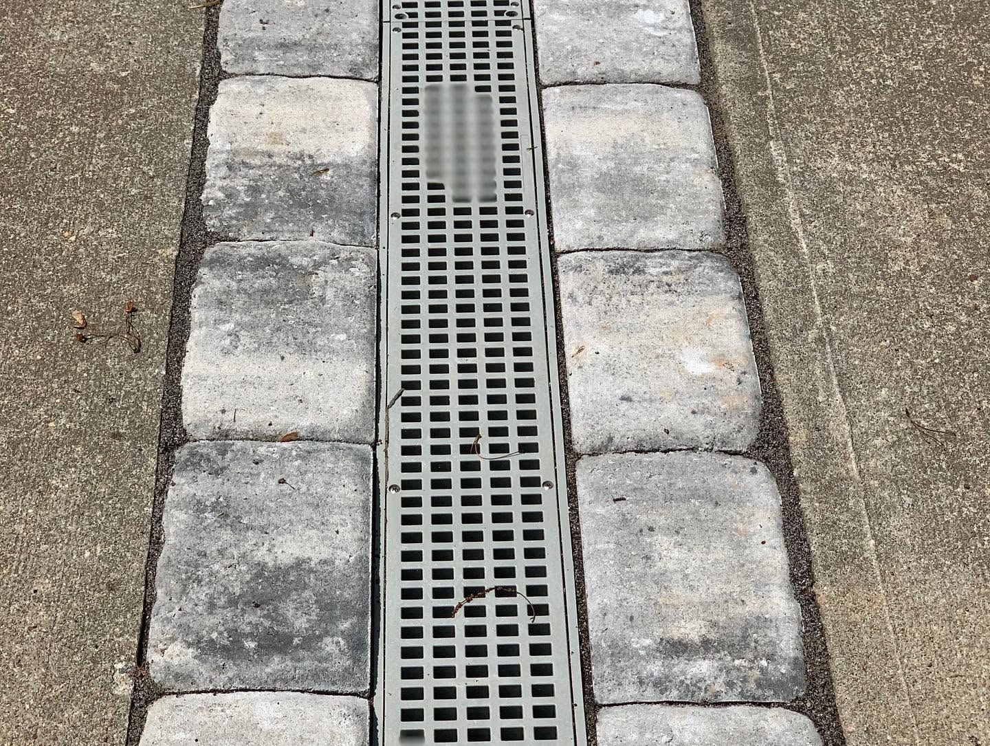 Narrow metal drainage grate bordered by gray pavers set in a concrete walkway.
