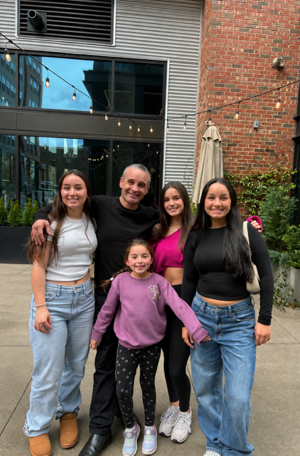Five people smiling outdoors in front of a brick building and glass storefront