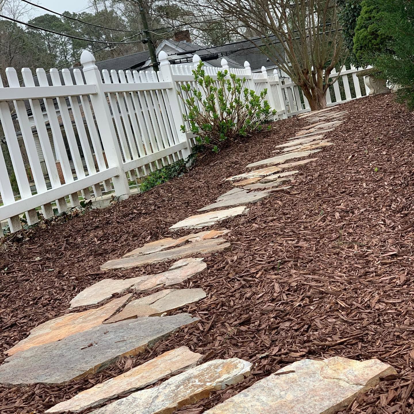 Stepping-stone path through a mulched yard beside a white picket fence and shrubs