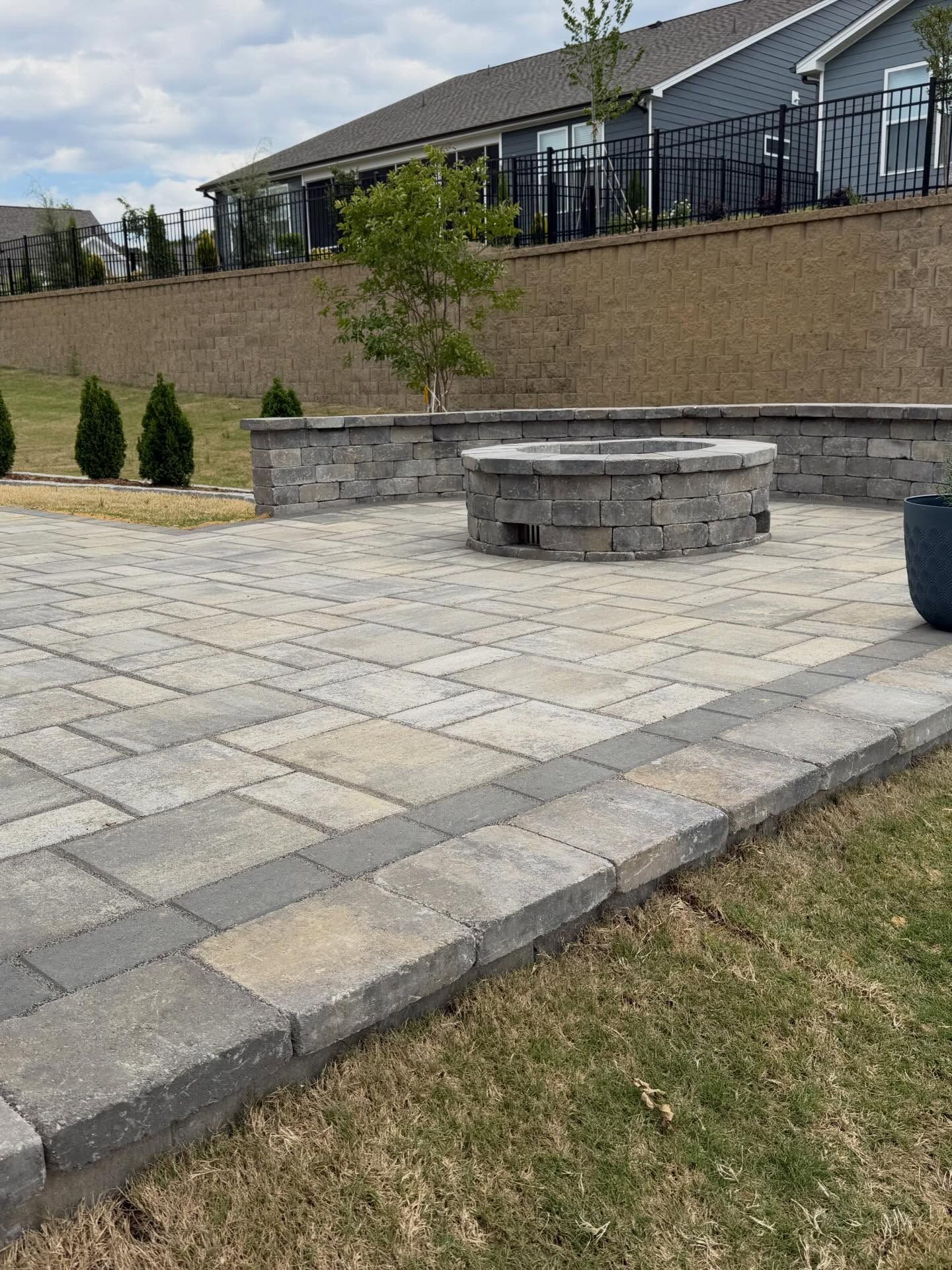 Stone patio with a circular fire pit and low retaining wall in a landscaped backyard.