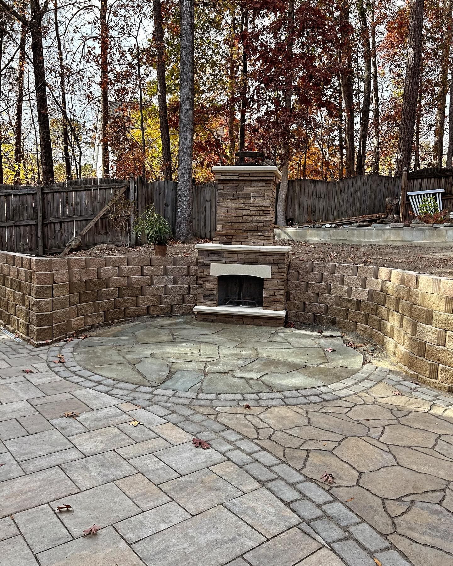 Stone patio with circular pavers, built-in outdoor fireplace, and low retaining walls in a wooded backyard