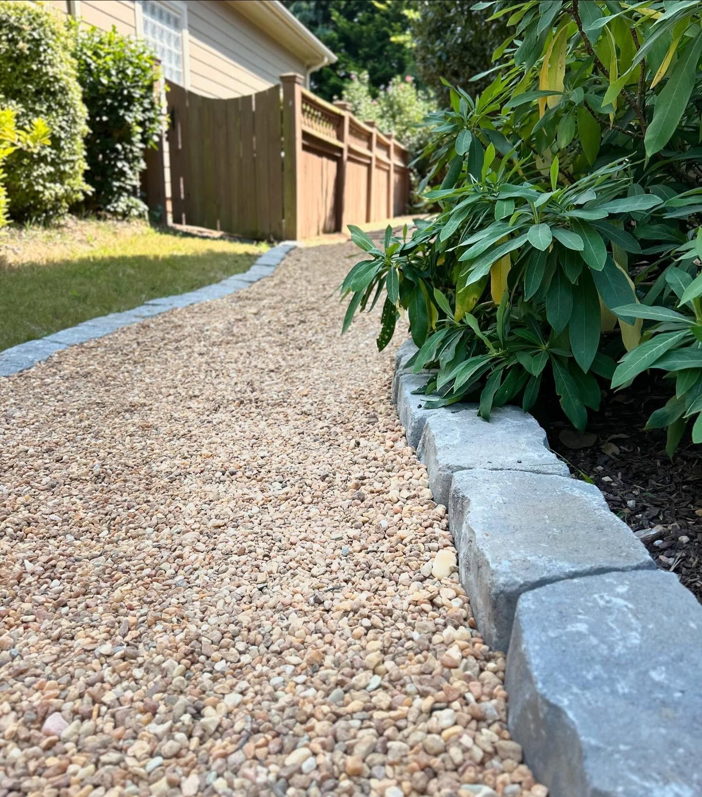 Pebble pathway beside a house, bordered by stone edging and green shrubs.