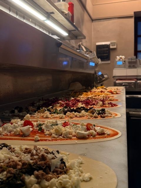 File di pizze crude con vari condimenti su un bancone da cucina sotto luci calde