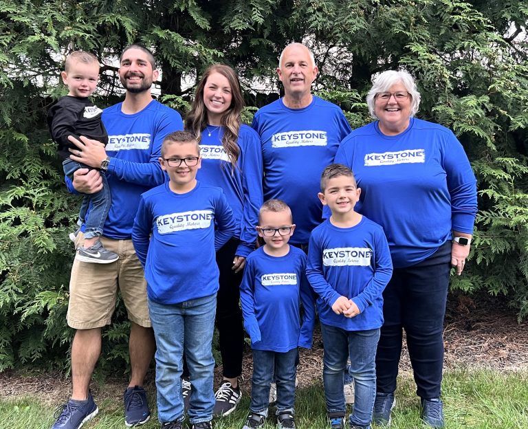 Seven people in matching blue Keystone shirts stand smiling outdoors in front of trees. | Keystone Quality Motors