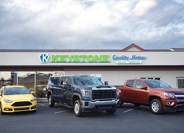 Car dealership lot with three parked trucks and a yellow car in front of Keystone logo signage | Keystone Quality Motors