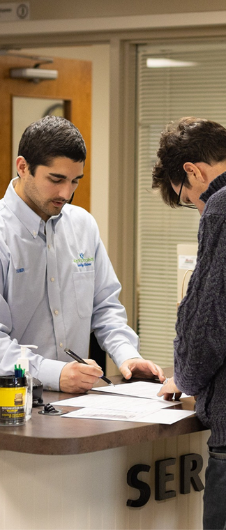 Two people at a service counter, one filling out paperwork behind a windowed office desk. | Keystone Quality Motors