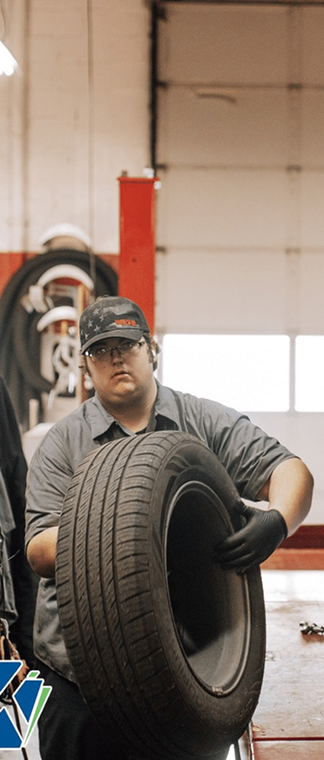 Mechanic in a garage lifting a large tire, with cars and shop equipment in the background | Keystone Quality Motors