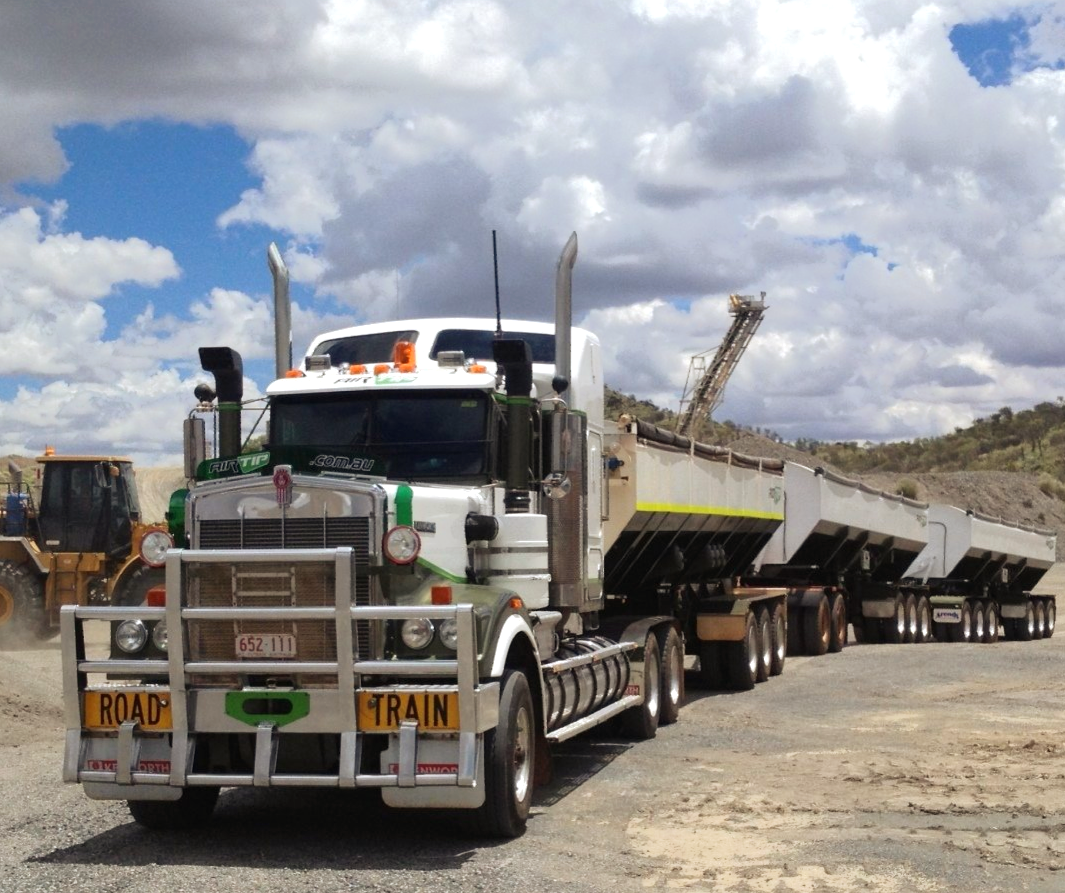 A white semi-truck hauling multiple trailers on a dirt road in a quarry under a cloudy sky.