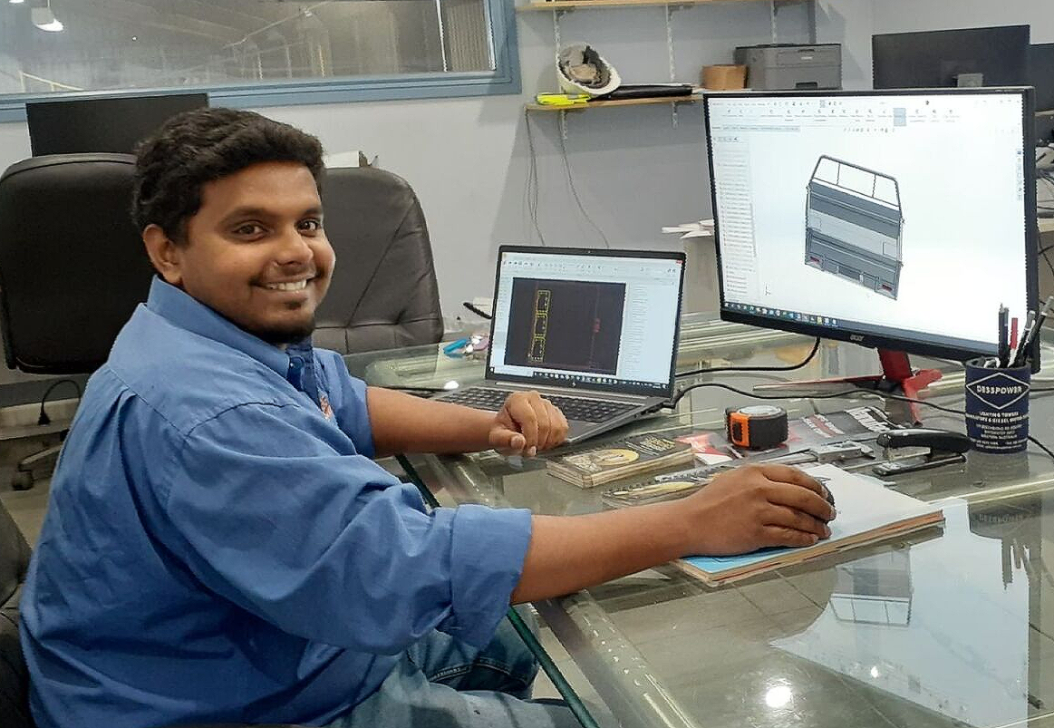 A man with dark hair and a blue shirt smiles while working on a computer with design software, indoors at a desk.