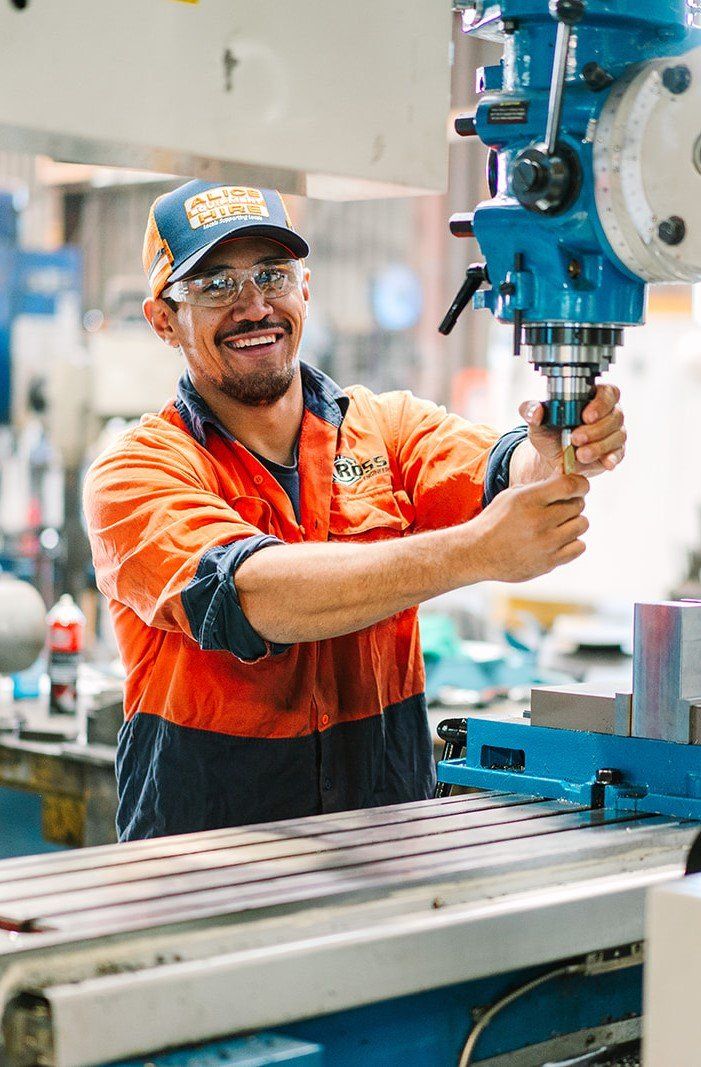 Smiling man in orange work shirt and cap operates a blue milling machine in a factory.