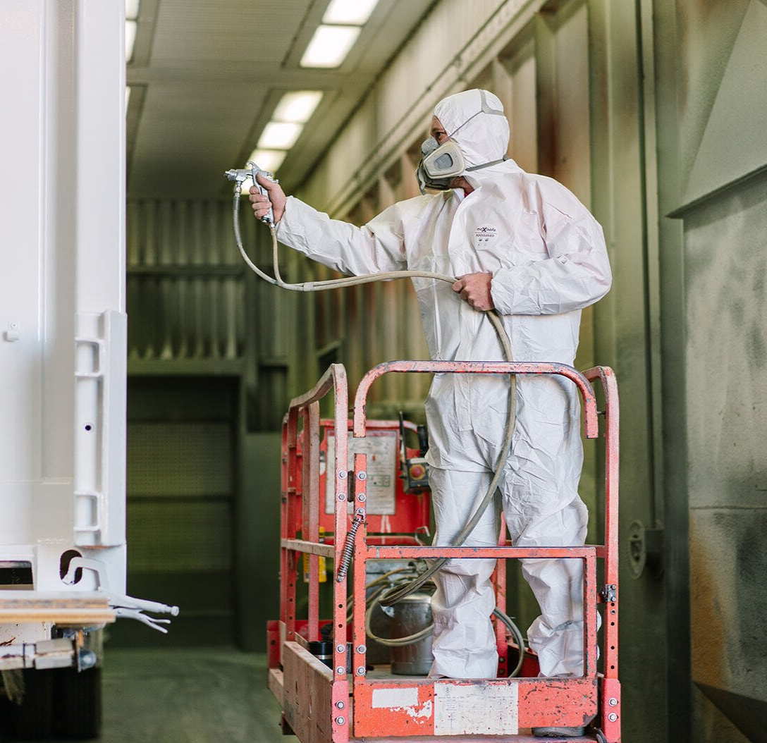 A person in a protective suit sprays paint on a large object while standing on a lift inside a paint booth.