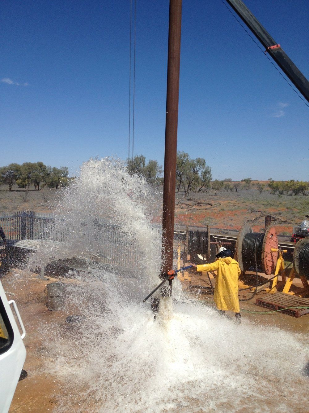 Water gushing from a well being drilled. A person in a yellow rain suit aims a hose as it sprays. Outdoors, sunny day.