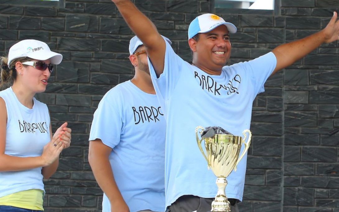 Three people in white shirts celebrate with a trophy, smiling and raising their arms outdoors.