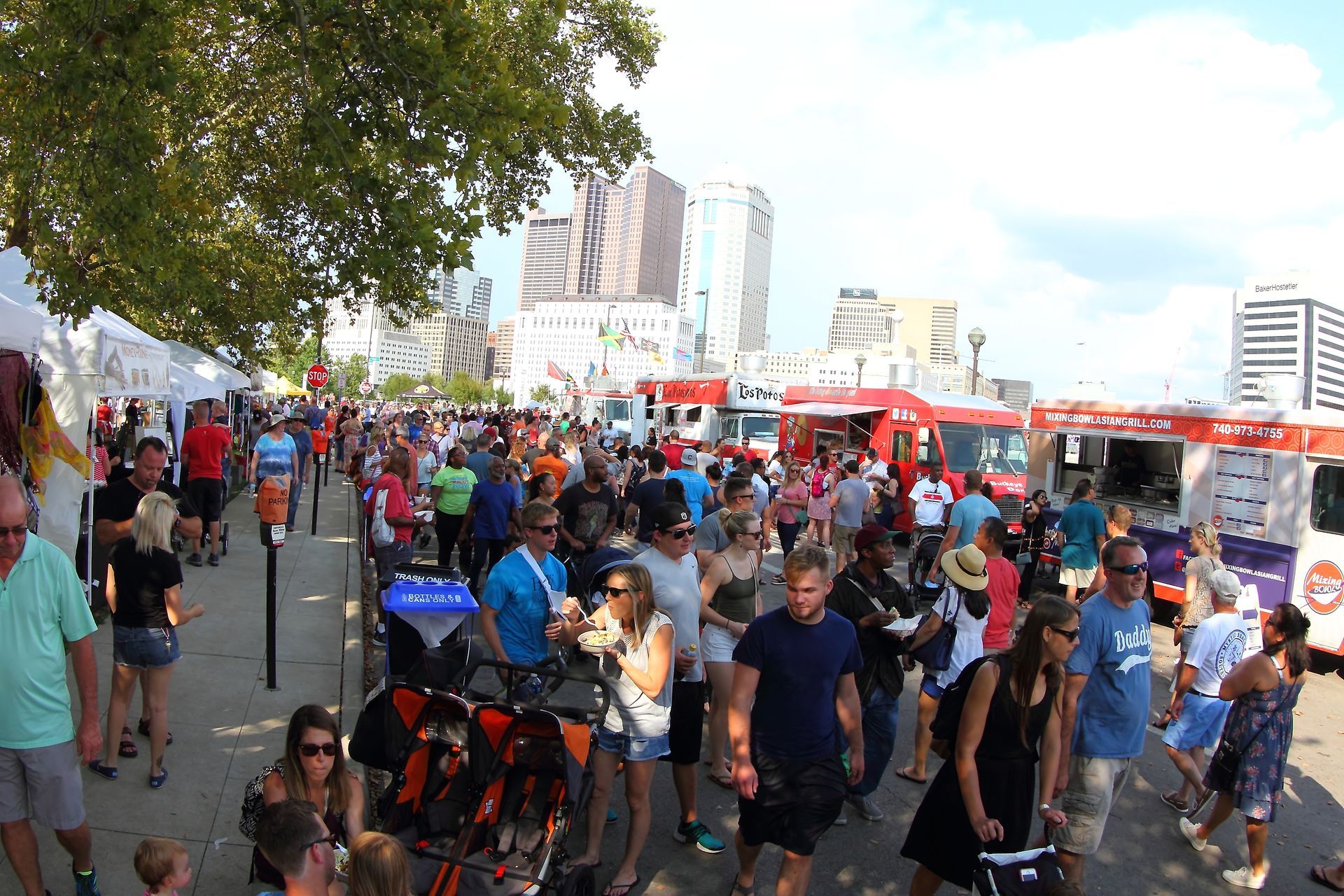 Crowded outdoor festival walkway with people, tents, and red vehicles under trees and city buildings