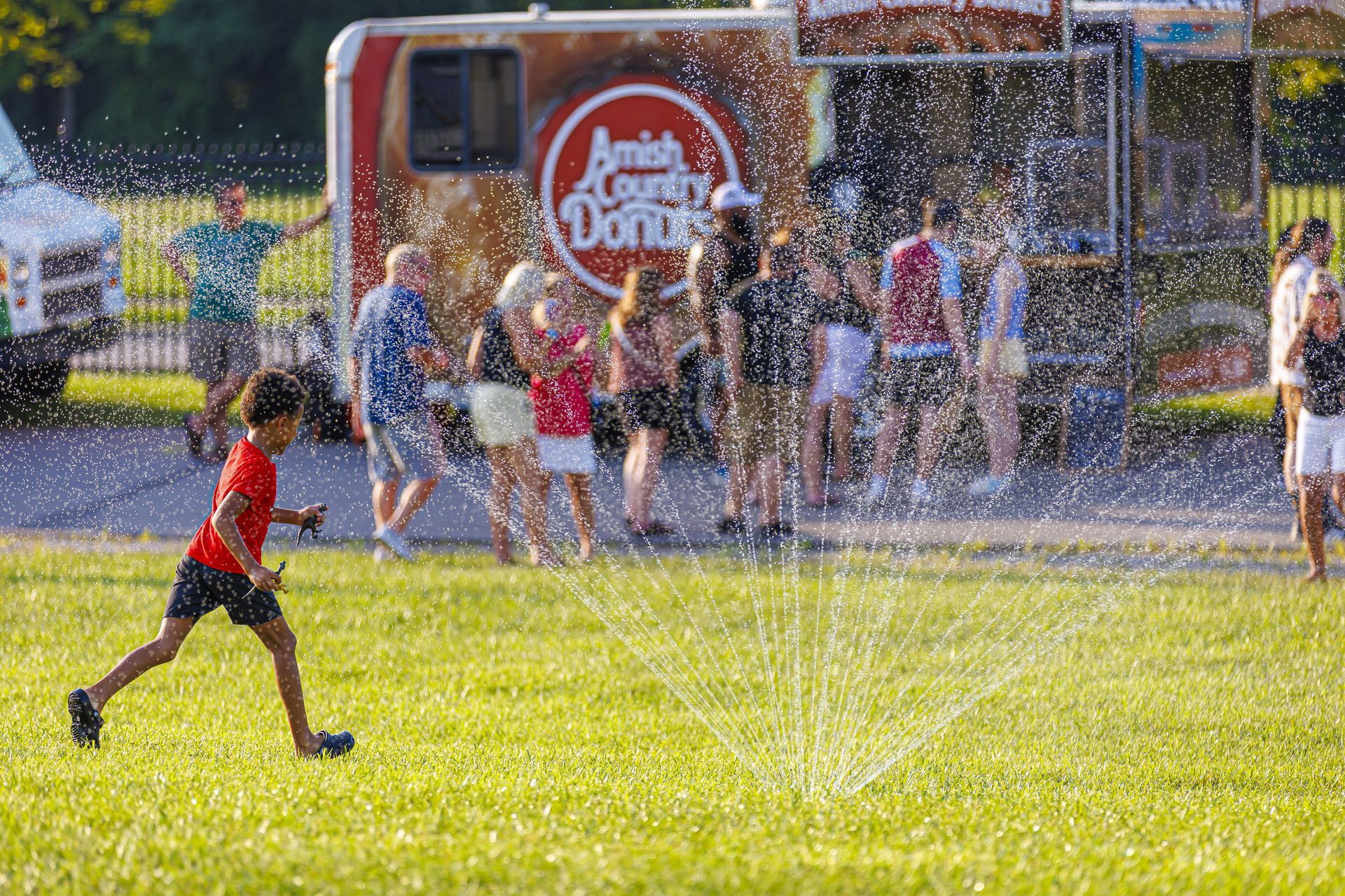 Child running through sprinkler on a grassy field at a fair, with people and an Amish Donuts truck in the background