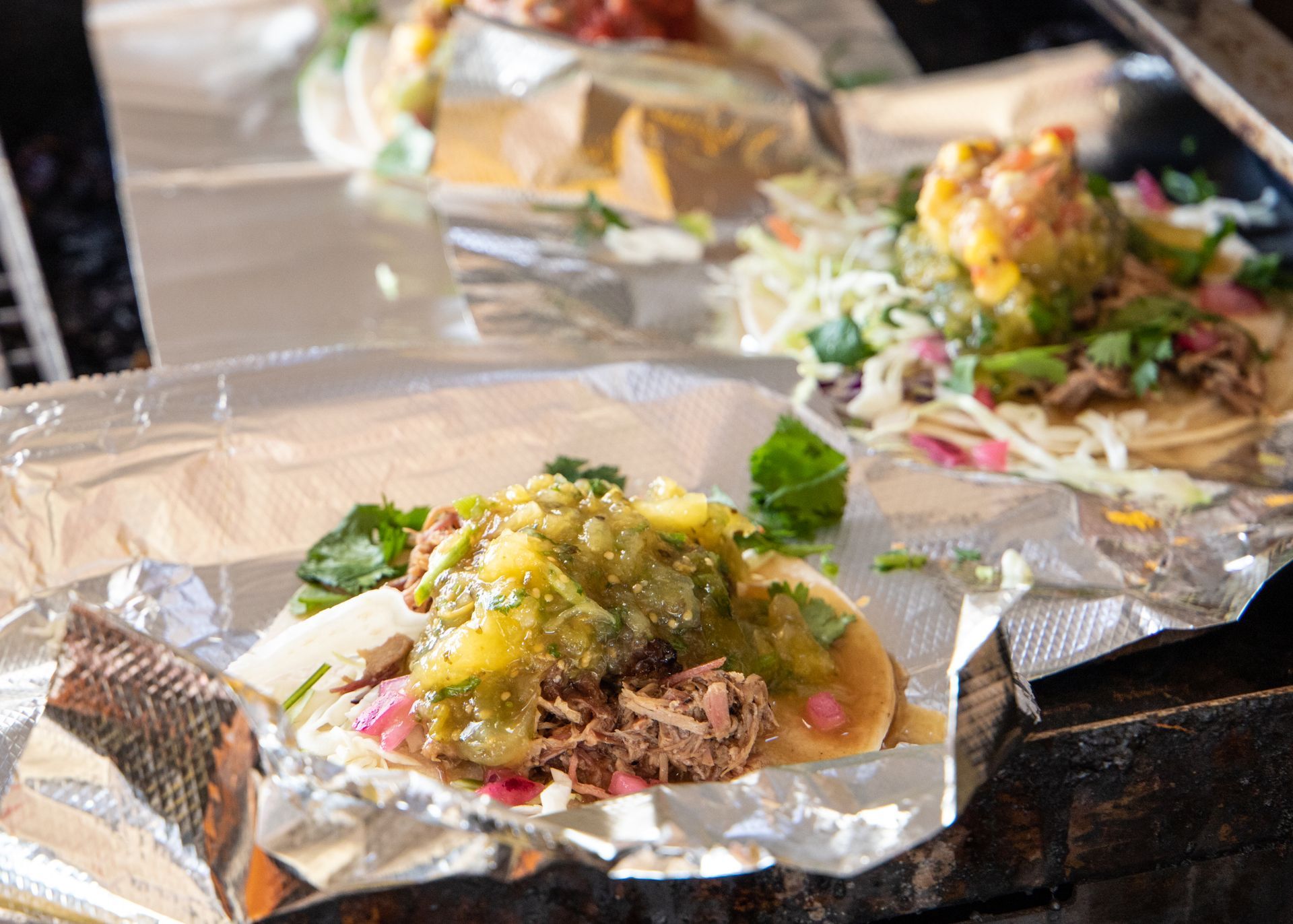 Tacos on foil-lined trays, topped with green salsa and garnishes, on a table
