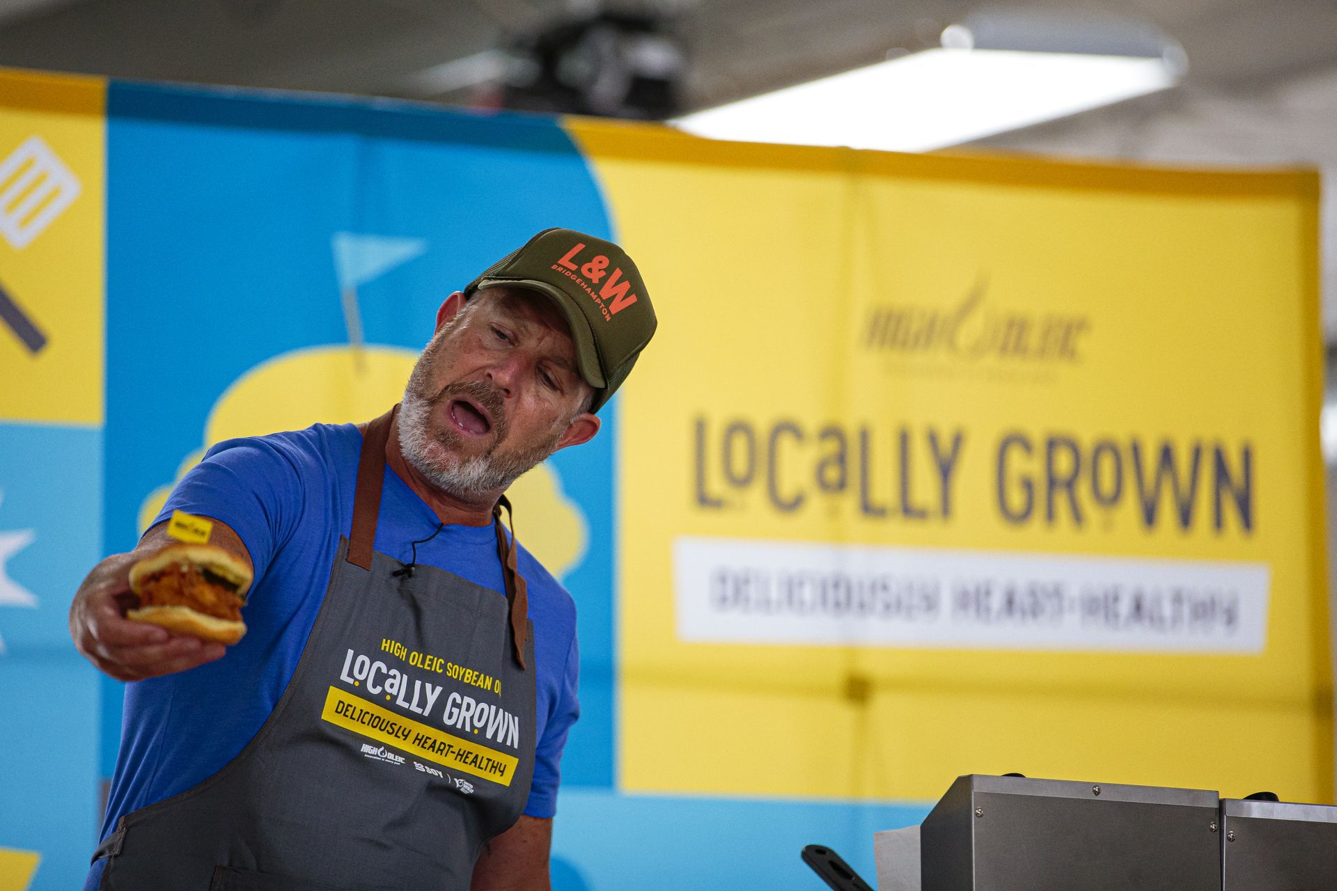 Man in apron and cap holding a burger onstage beside a “LOCALLY GROWN” sign