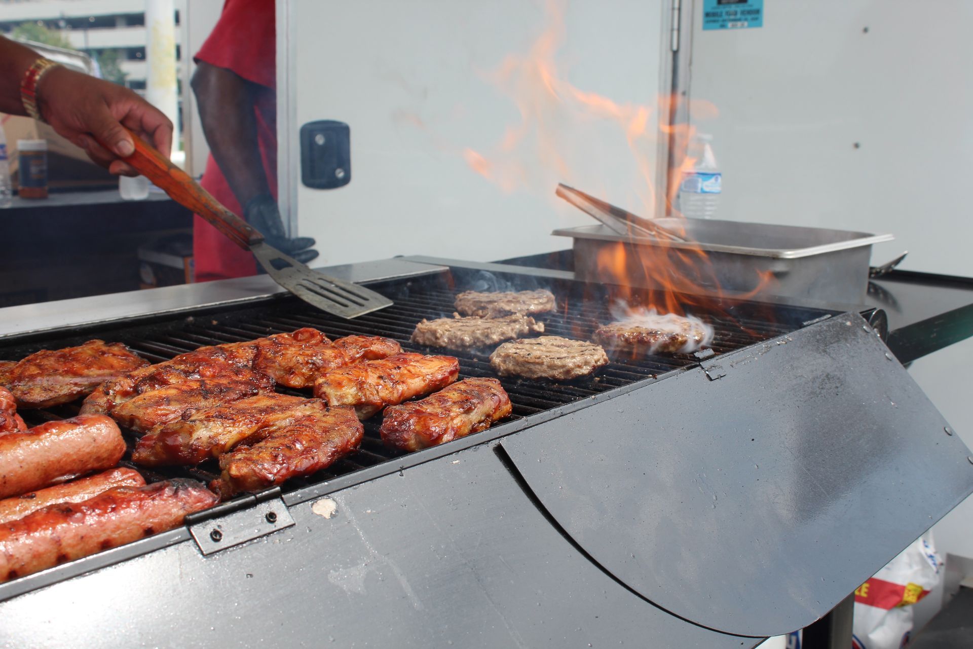 Person grilling sausages and burgers on a smoky outdoor barbecue.