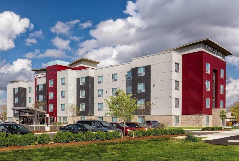 Modern apartment complex with red and white facades under a cloudy blue sky, with parked cars and green landscaping.