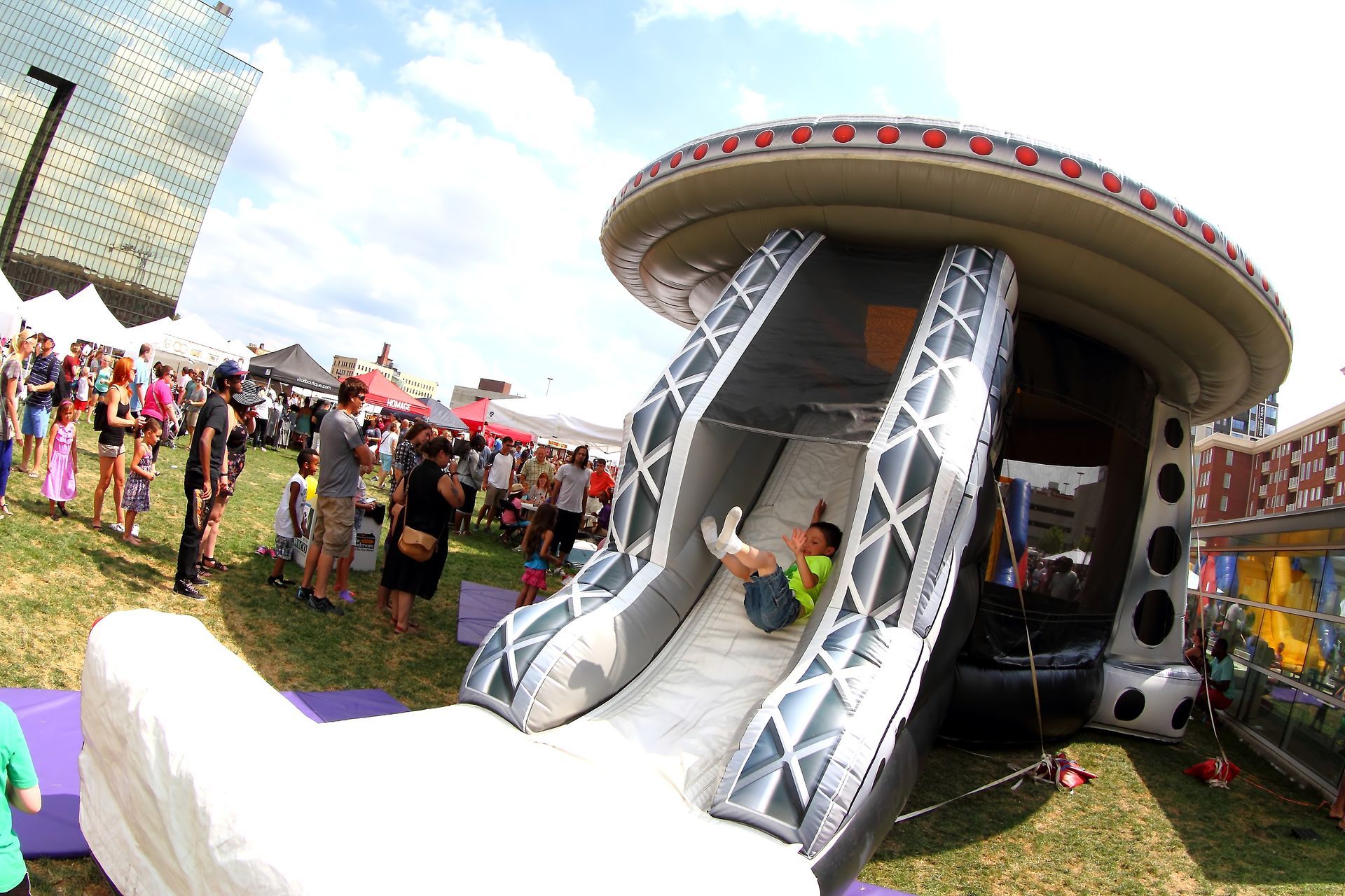 Crowd at an outdoor event with a large inflatable slide and a round play structure on a sunny day