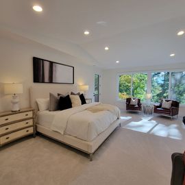 Spacious neutral bedroom with a large bed, dresser, sitting area, and bright windows overlooking greenery
