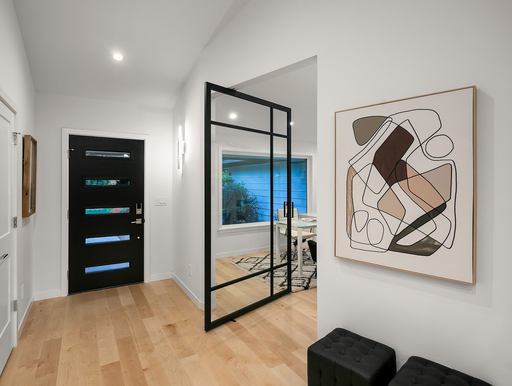 Modern hallway with wood floor, black-framed glass door, abstract wall art, and view of a blue-lit room.