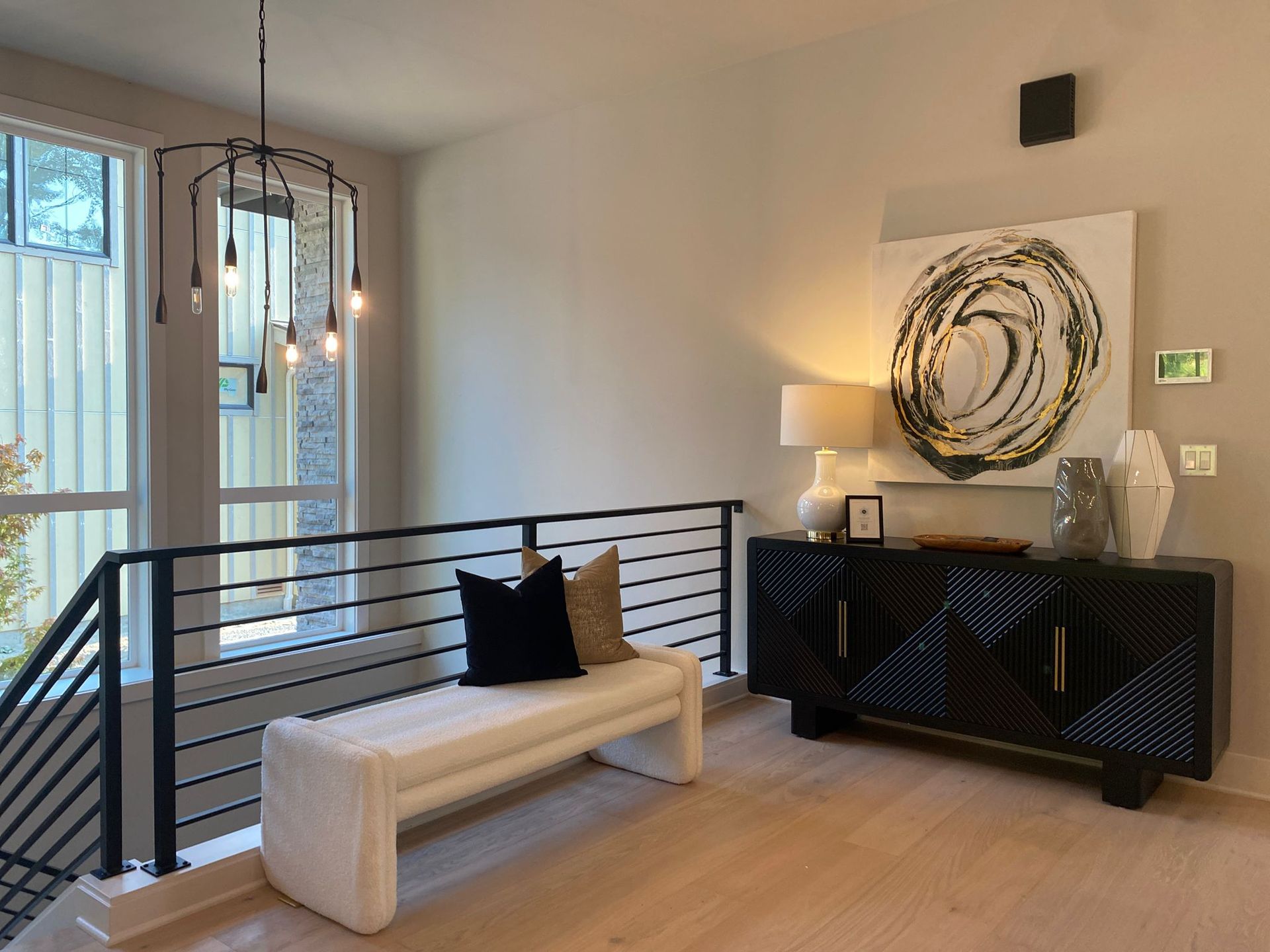 Modern living room with beige bench, black railings, chandelier, and dark sideboard beneath abstract wall art