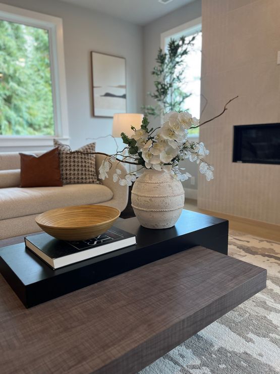 Modern living room with a black coffee table, white vase of flowers, and neutral sofa near a window.
