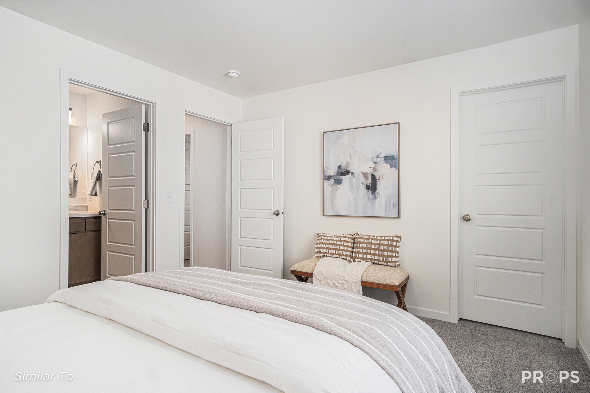 Bright white bedroom with bed, bench, artwork, open bathroom door, and closet doors
