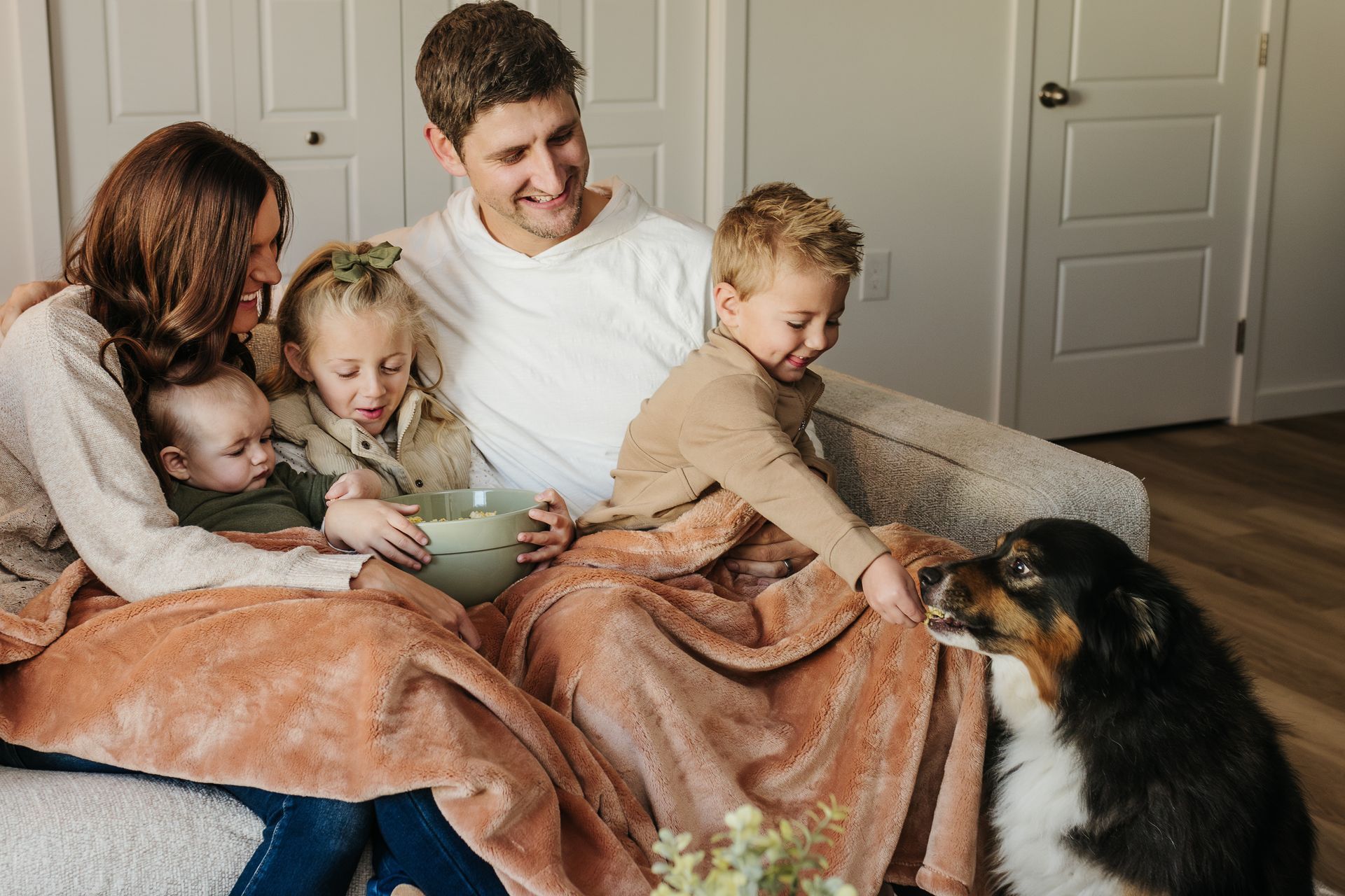 Family on a couch shares a blanket and treats a dog indoors on a cozy afternoon
