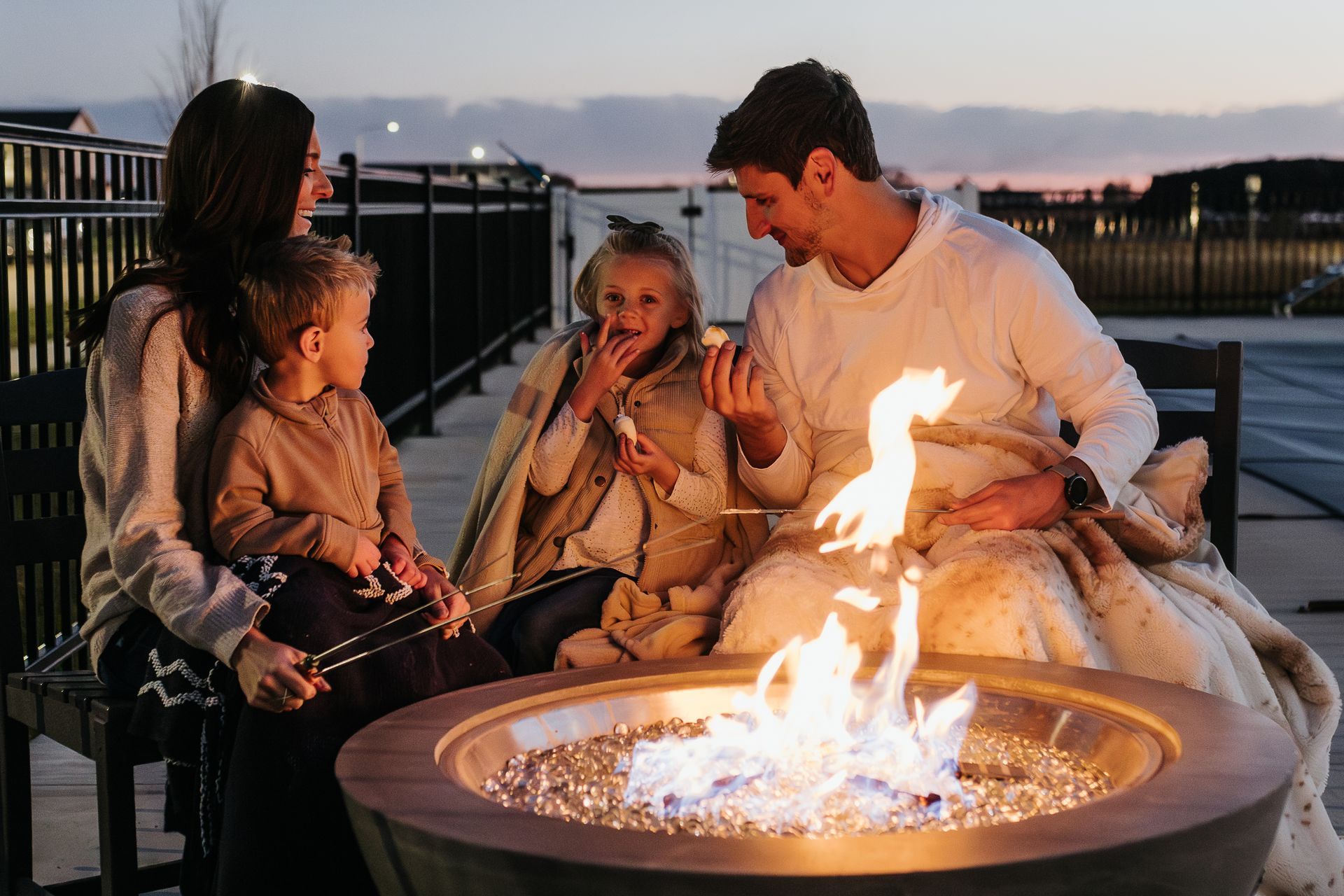 Family sitting around a fire pit outdoors at dusk, warming hands and talking on a patio.