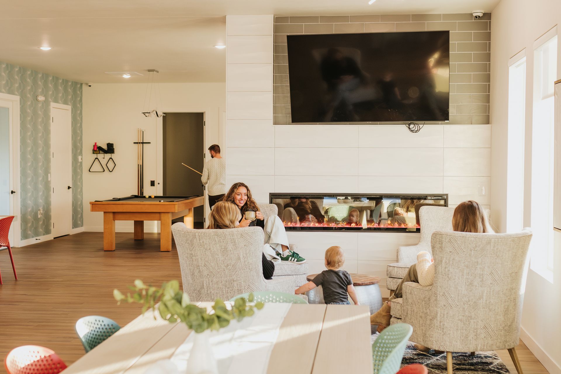 Bright living room with people seated near a fireplace and a large wall-mounted TV