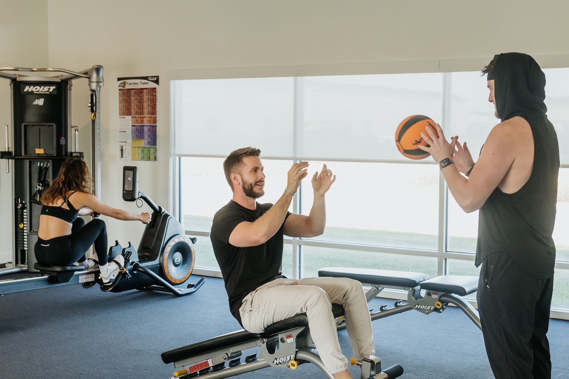 People exercising in a gym, with one holding a medicine ball while another claps on a rowing machine.
