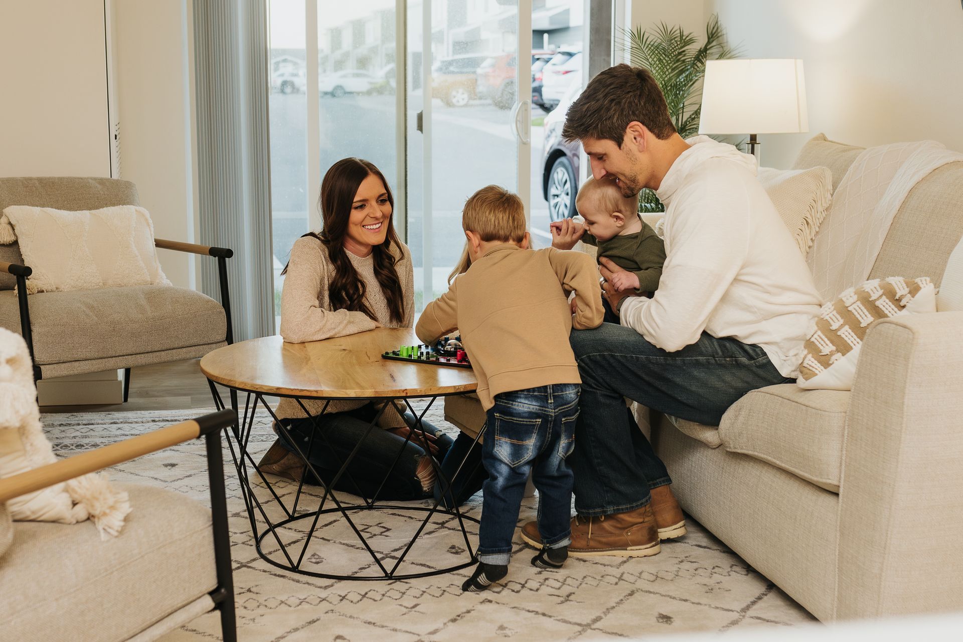Family gathered around a coffee table in a bright living room, sharing a playful moment with a toddler.