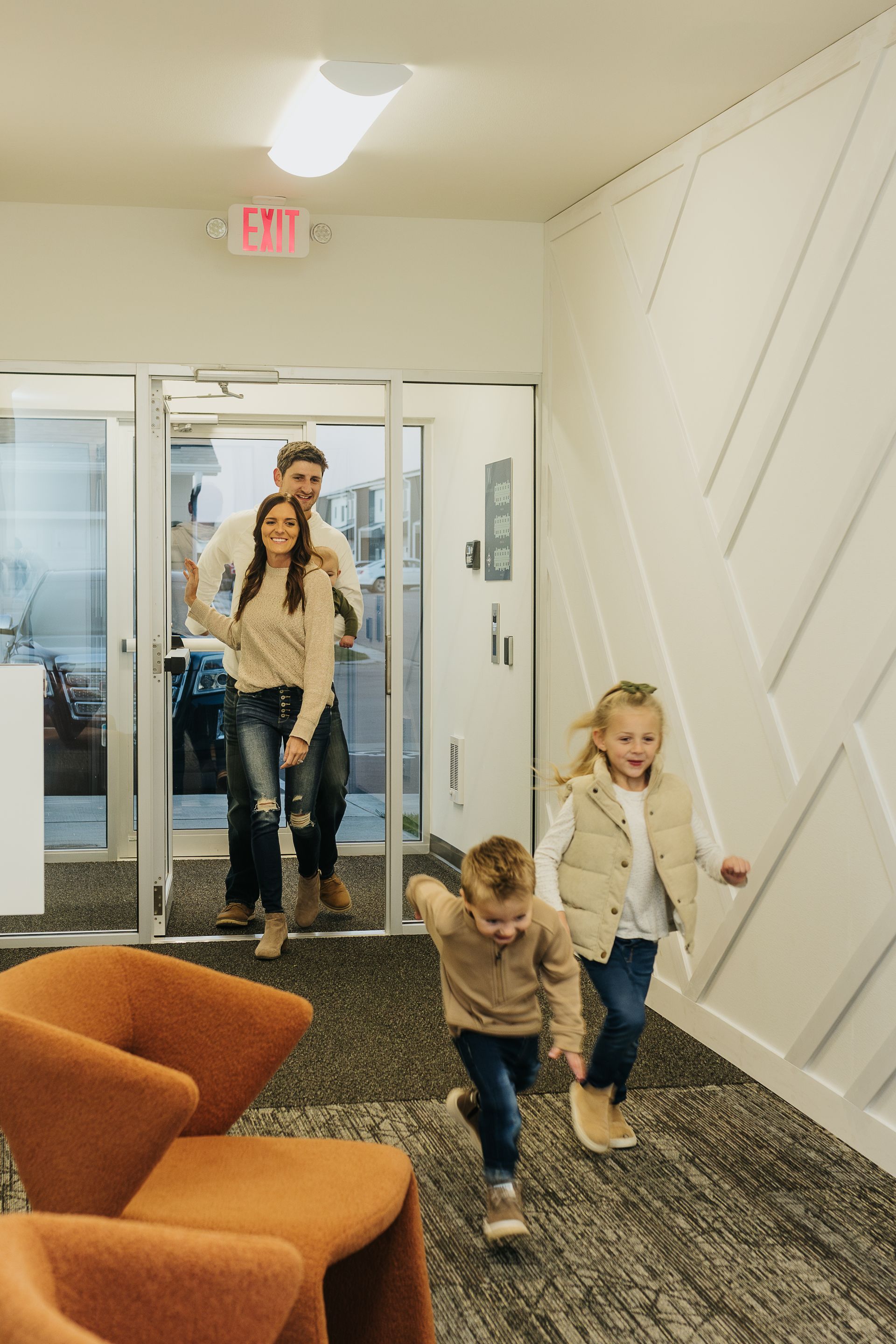 Two children run through a carpeted office lobby while a woman stands by the glass entrance.
