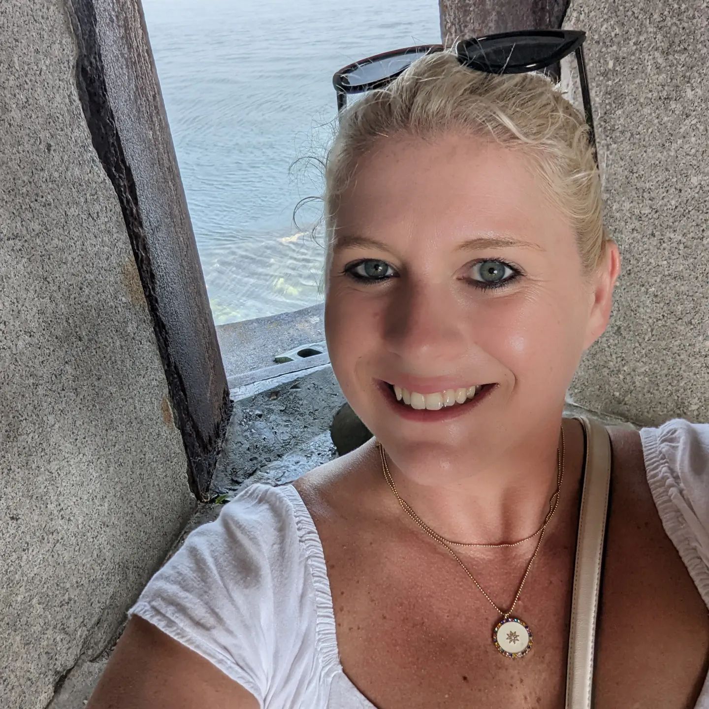 Smiling blonde woman taking a selfie by the water, wearing sunglasses on her head and a white top.