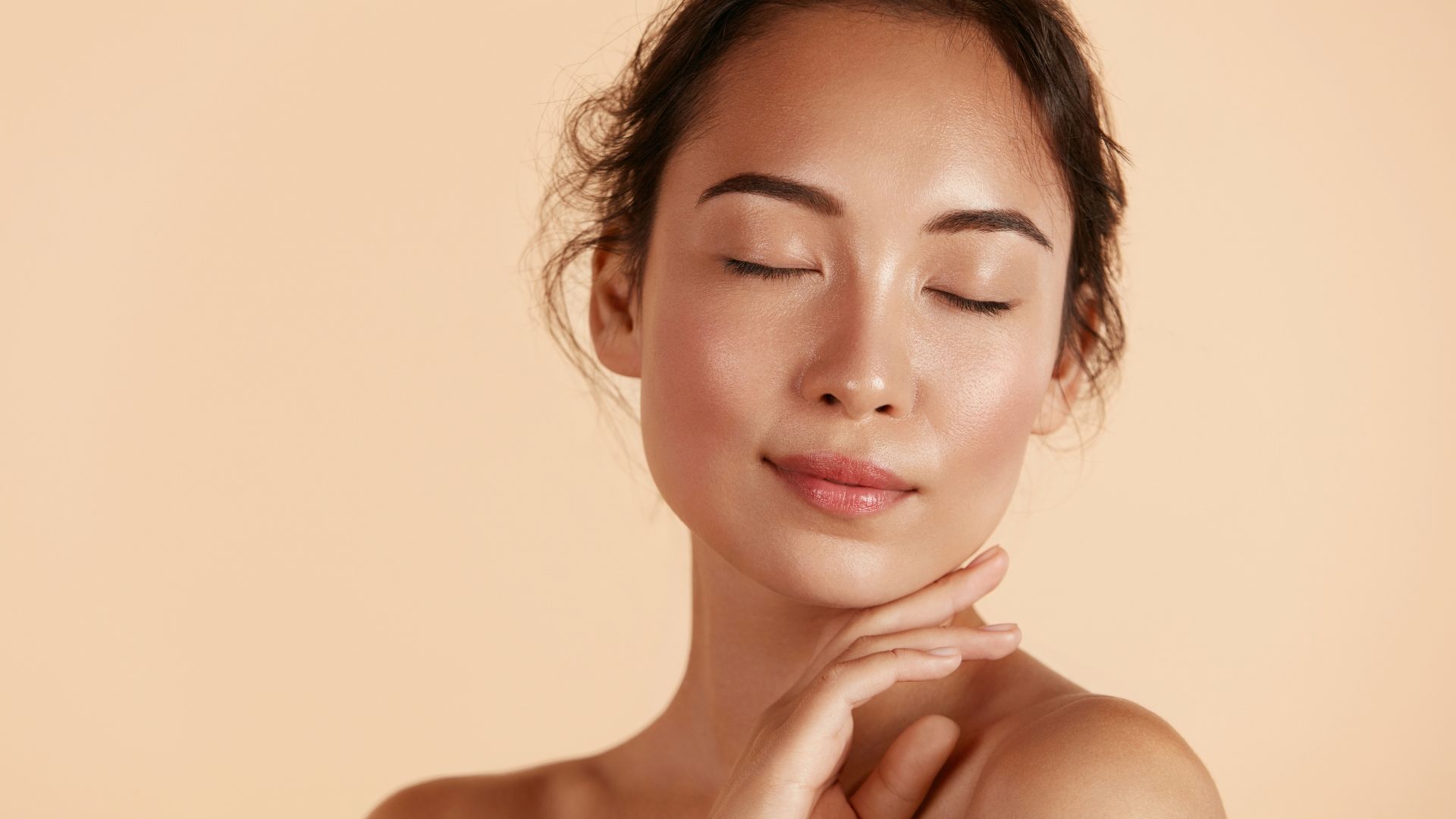 Close-up of a person with eyes closed, gently touching their neck against a soft beige background