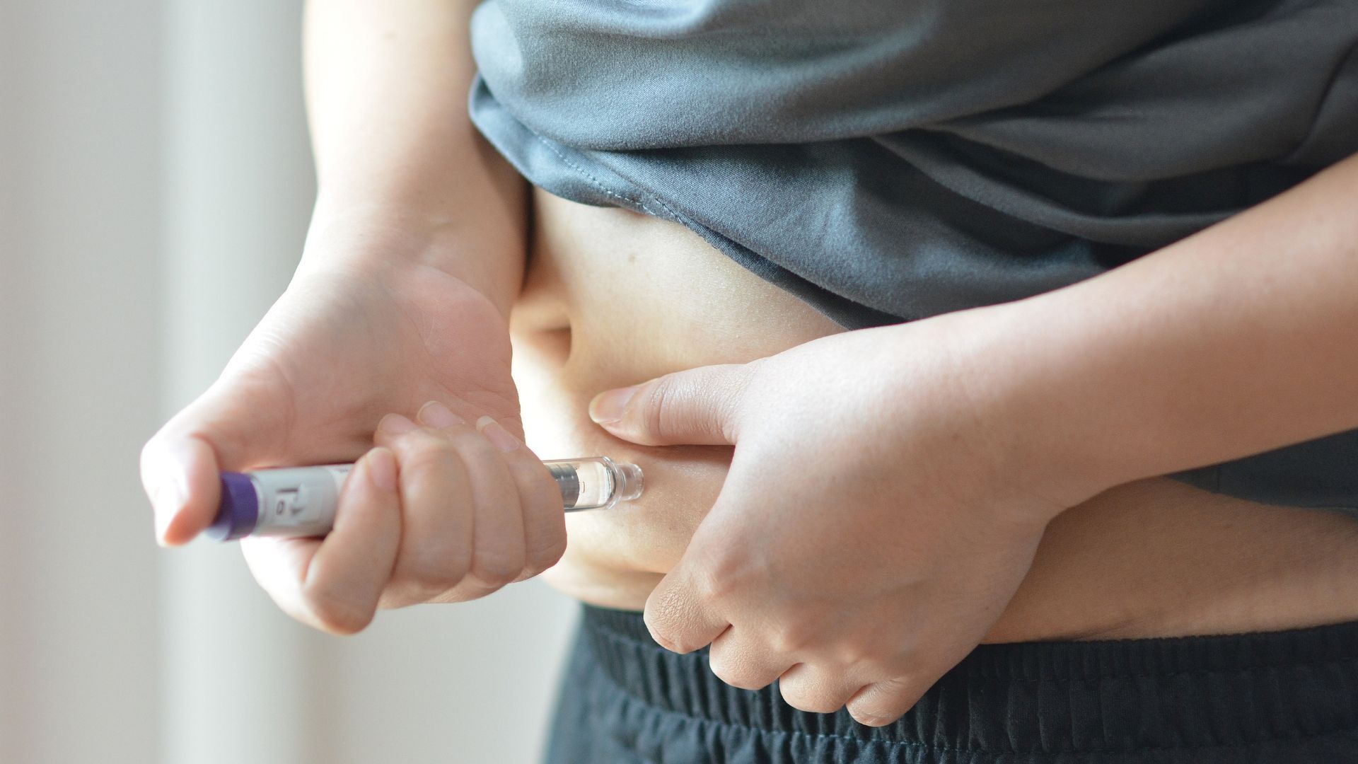 Person injecting medication into abdomen with a syringe, wearing a gray shirt and black pants.