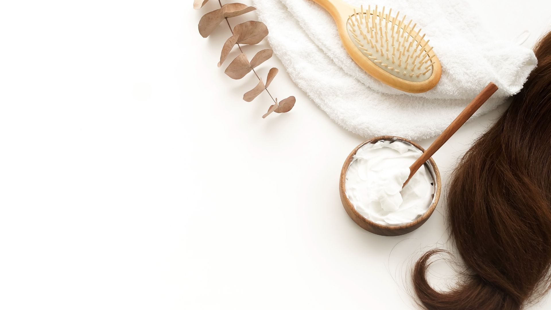 Hair care flat lay with coconut bowl, wooden comb, dried leaves, and brown hair on a white background